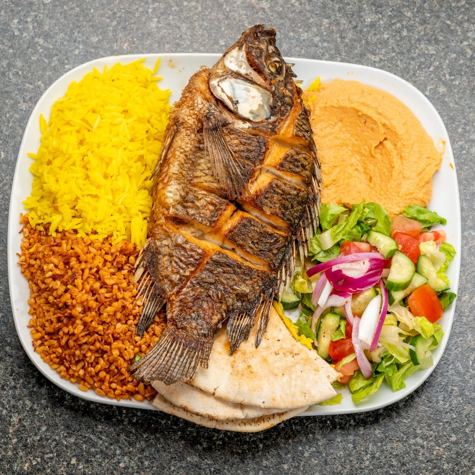 A whole fried fish with yellow rice, bulgur, hummus, salad, and pita bread on a white plate.